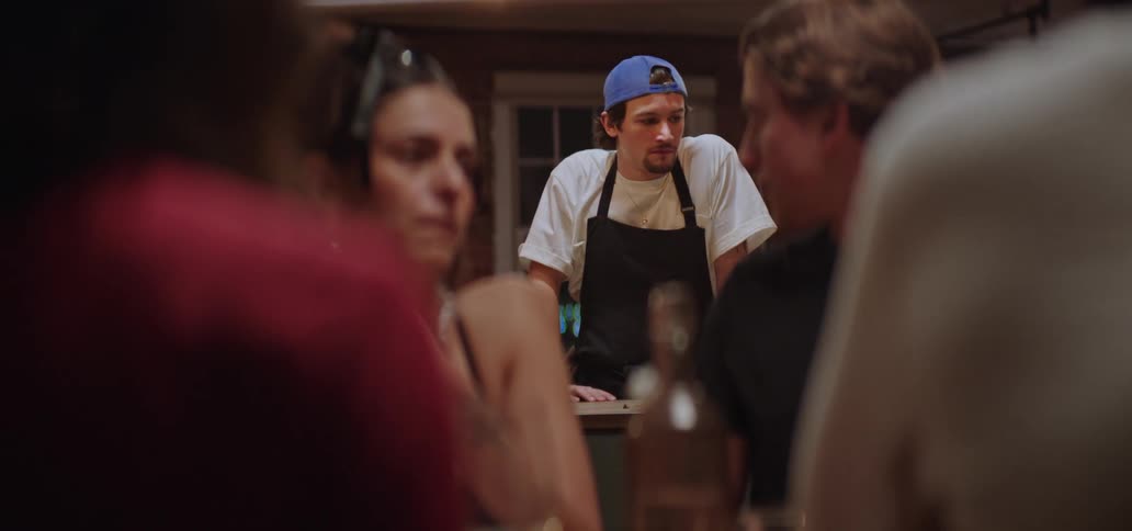 A man in an apron leans on a kitchen counter as people converse nearby