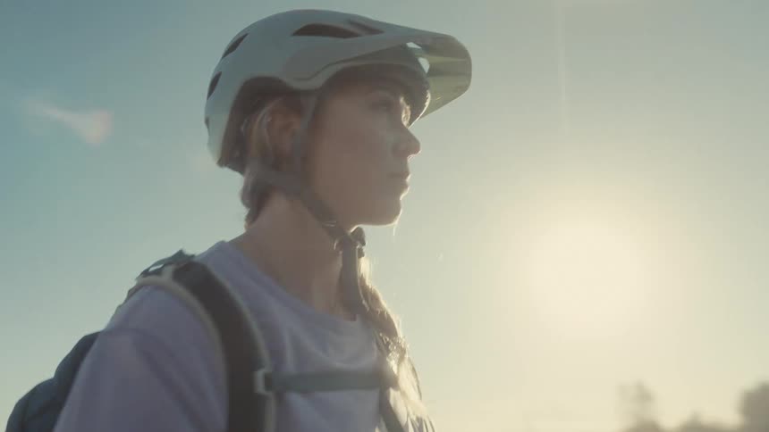 A cyclist wearing a light blue helmet and backpack is captured in closeup backlit by the sun she looks thoughtfully across a sunny open landscape with trees in the distance