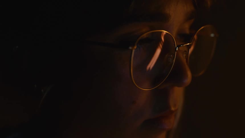 A close up of a persons face who is in dim light looking forward wearing eyeglasses