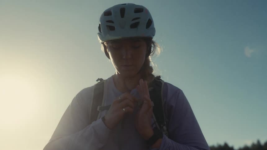 A female cyclist wearing a helmet and backpack adjusts her gear against a bright sunlit clear sky background she appears focused and ready as she looks offcamera into the distance