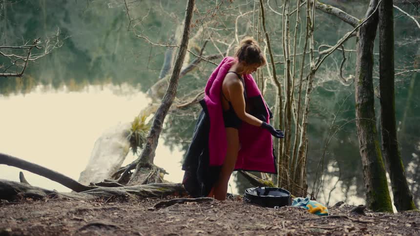 Woman wrapped in a pink coat picking up items beside the lake