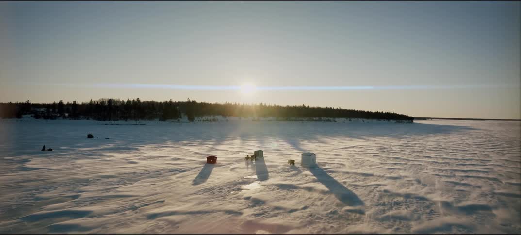 An aerial wide shot captures several small fishing shacks on a vast snow covered frozen lake near a dark forest under a bright sun