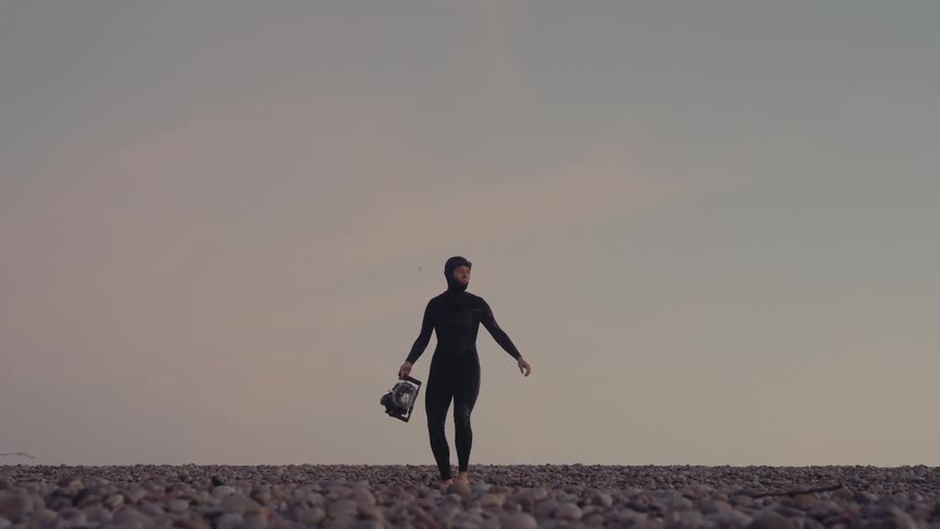 The video features a person in a black wetsuit walking across a pebble beachthe person is holding what appears to be a camera housing or a similar piece of equipment possibly preparing for an activity like underwater photography or diving