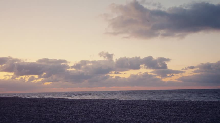 Clouds fill the sky above a pebble beach and calm ocean at sunset