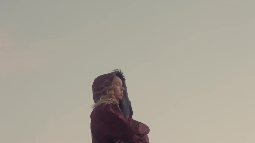 A young woman in a hooded jacket stands with crossed arms against a pale sky