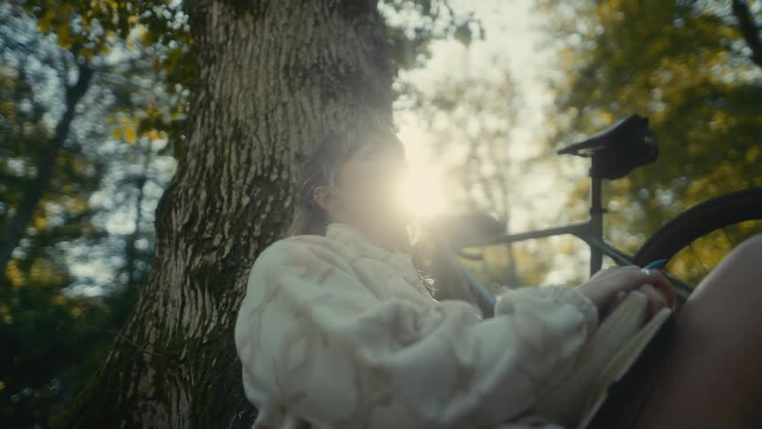 Teenage girl sits leaning against a tree in the forest next to her bicycle as the sun shines brightly in the distance