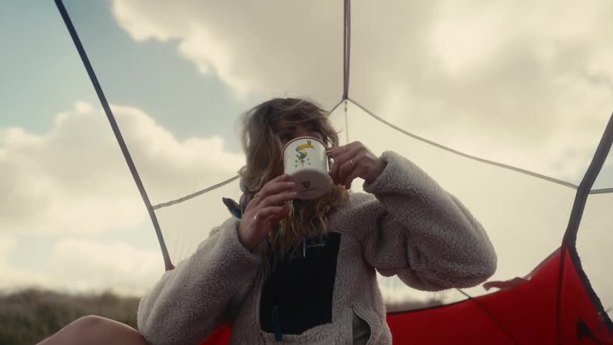 A young woman in a fleece jacket sips from a mug inside a tent