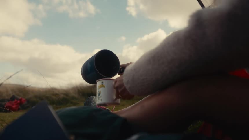 A person is sitting outdoors pouring coffee from a blue jug into a white mug