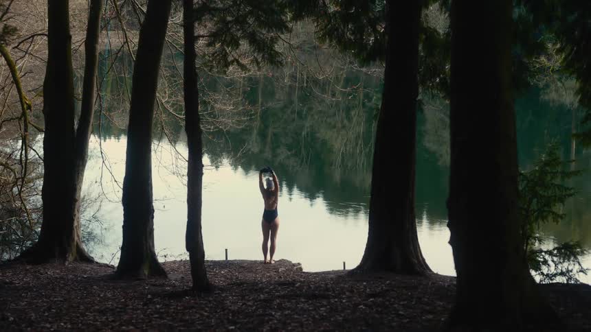 A young woman in a swimsuit stretching on the bank of a lake surrounded by tall trees