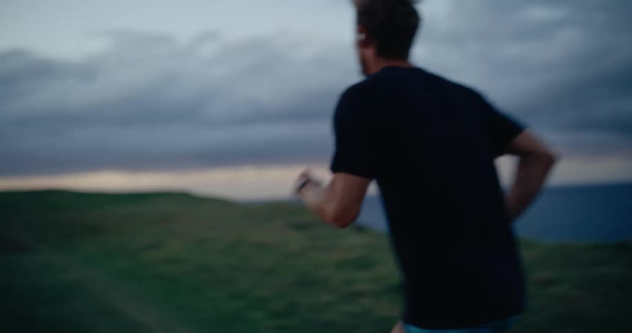 A man jogs along a grassy coastal cliff overlooking the ocean at dusk