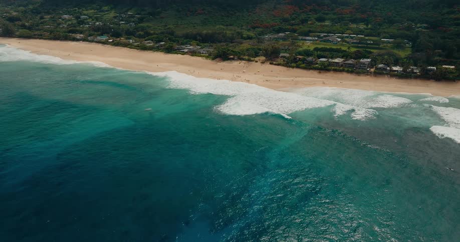 An aerial shot overlooks a tropical coastline where turquoise waves crash onto a sandy beach lined with lush greenery and homes