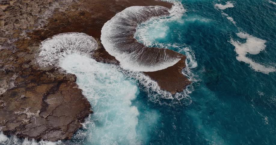 Aerial view of powerful ocean waves crashing over a rugged dark brown rock shelf creating white foaming water that flows back into the deep blue sea