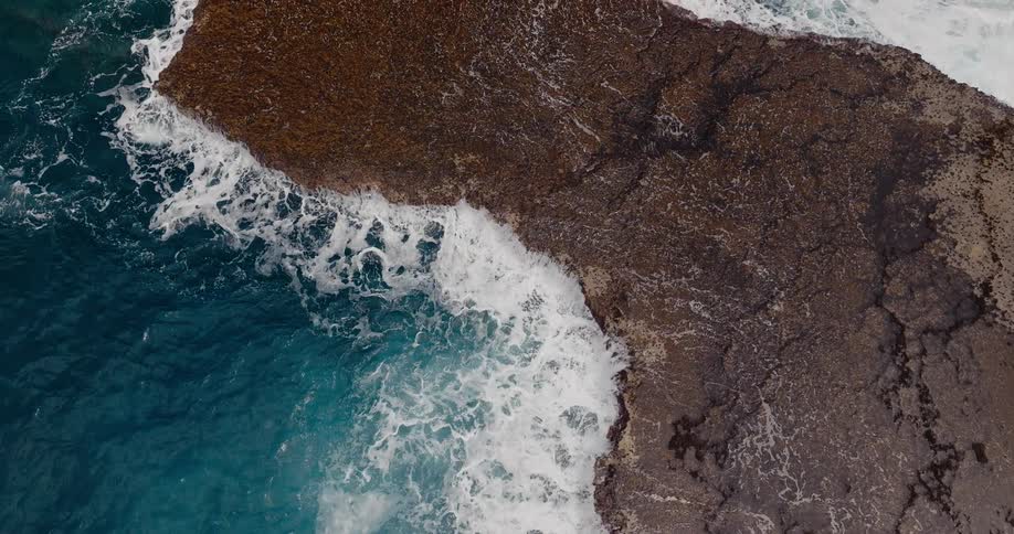 Overhead aerial view captures deep blue ocean waves crashing against dark jagged rocks creating white foam and swirling water patterns