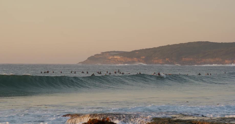 A wide shot captures many surfers waiting in the ocean water for waves near a sandy beach as gentle white waves roll toward the shore