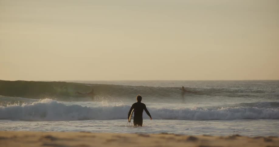 Male surfer carrying a surfboard and walking towards the sea where other people are surfing outdoors during day time