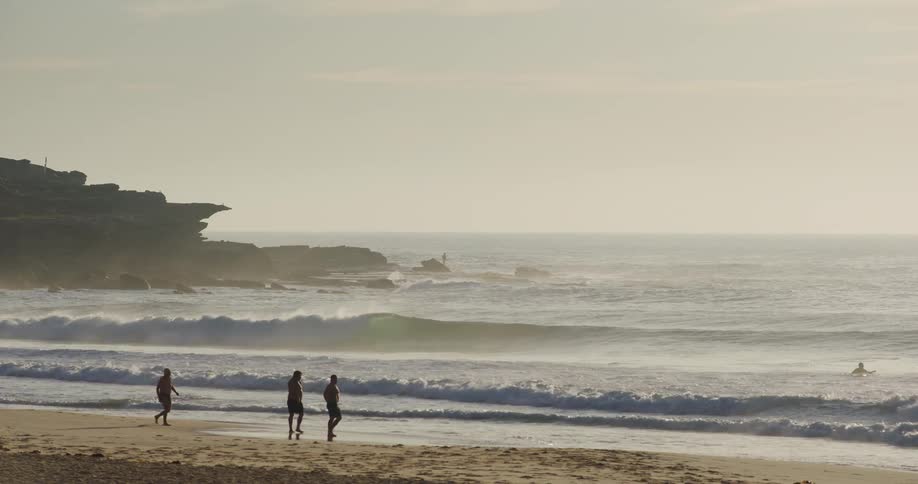 Men wearing dark shorts and no shirts walk along a sandy beach at sunset as large ocean waves crash near a rocky cliffside