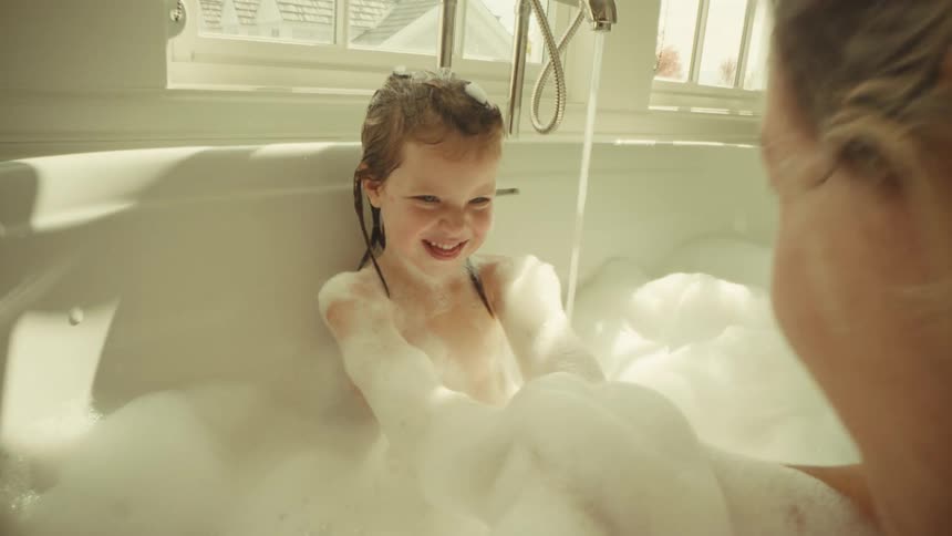 A happy little girl with wet hair plays in a bubble bath smiling at the camera as a woman bathes her near a bright window
