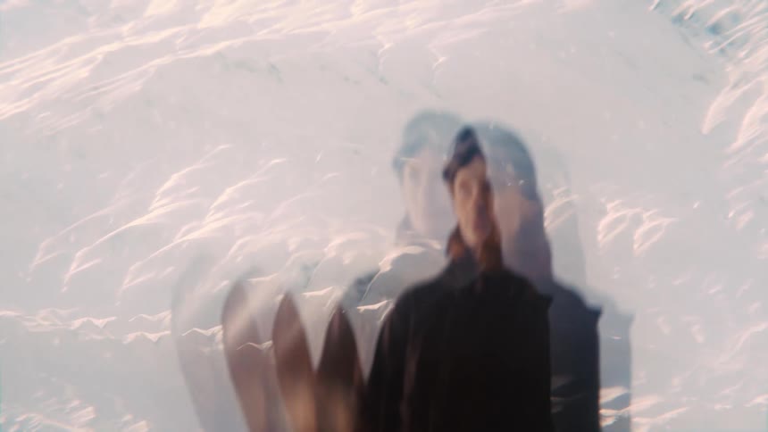 A dreamy layered visual shows a woman holding her snowboard her ghostlike figure blending into the bright sunlit snow of a vast mountain