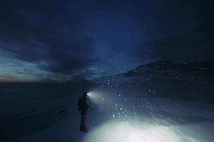 Person stands on a snow covered area with a headlight illuminating the ground nearby during a snowstorm