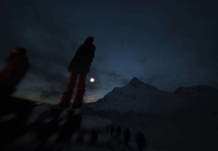 In a dark mountain landscape two people stand on the roof of an offroad vehicle near a small campfire gazing at a large mountain peak beneath the rising moon