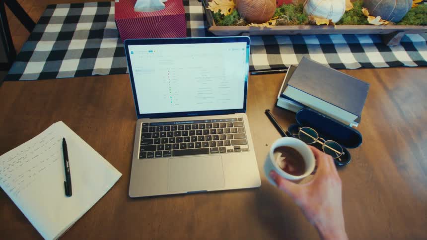 Person typing on a laptop while consulting notes and drinking coffee