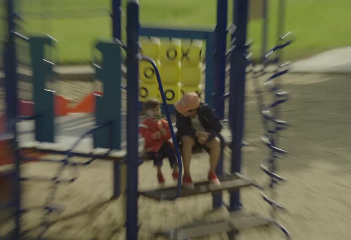 This video shows two children sitting on a structure at a playground on a sunny daythe younger child wearing a red and white jacket is licking a large green and white lollipop while the older child wearing a dark jacket and a sunny baseball cap