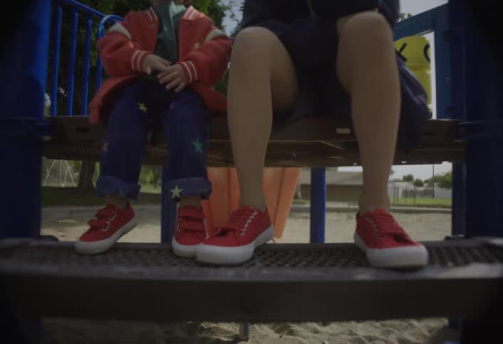 The video shows two children sitting sidebyside on a wooden beam of a blue playground structure filmed from a low angle the older boy who is on the right wears a dark jacket dark shorts and a red cap and he has his hand covering his mouth or chin