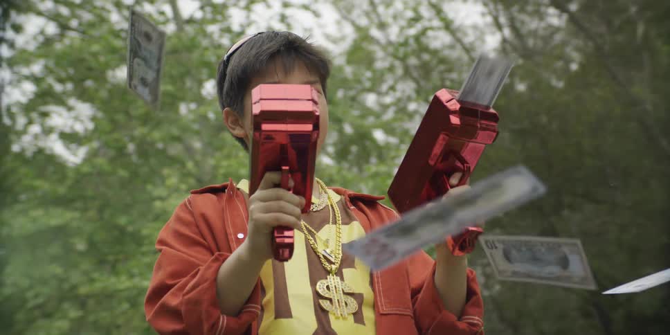 Young boy standing outside holding two red toy guns shooting out hundred dollar bills