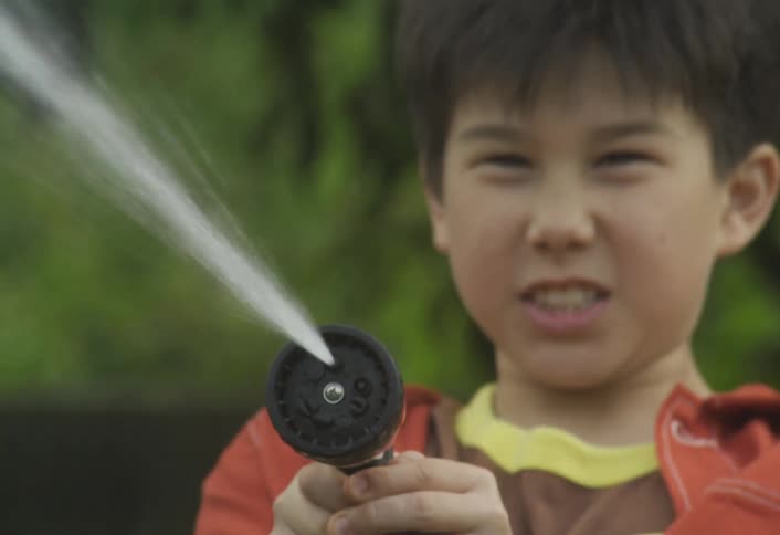 The face of a teenage boy holding a hose pointing it forward and spraying water while displaying a determined expression