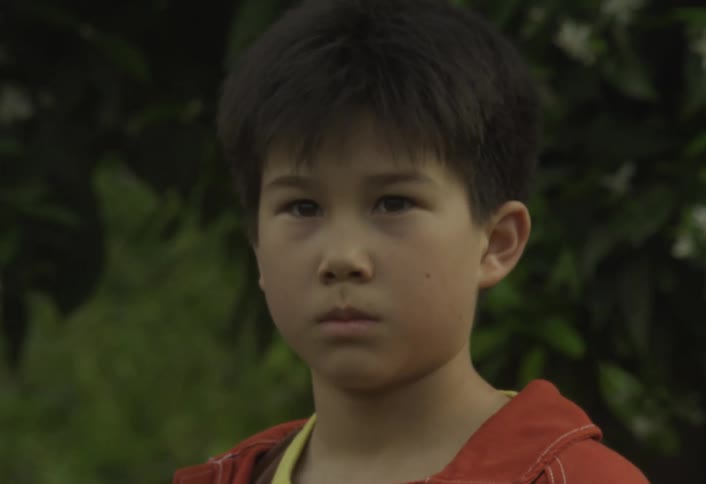 The camera zooms in on a young boys serious face which suddenly breaks into a playful grin as he aims and sprays a powerful stream of water from a hose