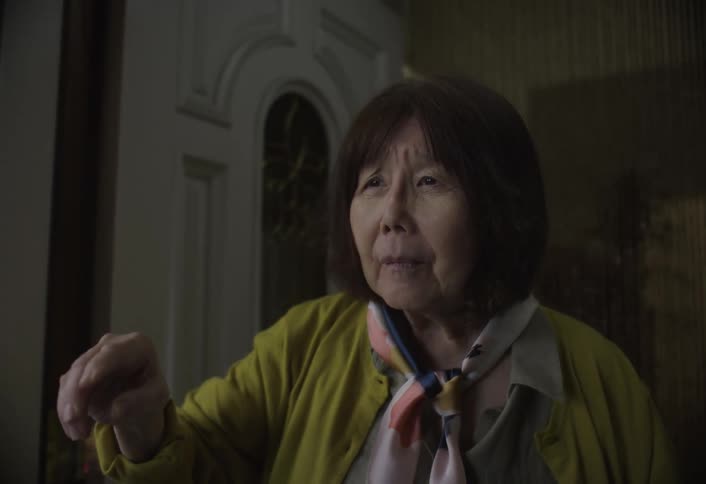 A closeup of an elderly woman with short dark hair wearing a yellow cardigan and a scarf standing in a dim doorway and actively speaking while raising her hand to gesture