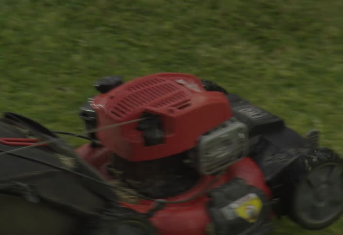 A young boy wearing a cap and green uniform pushes a red selfpropelled lawnmower across a bright green lawn in a closeup outdoor scene