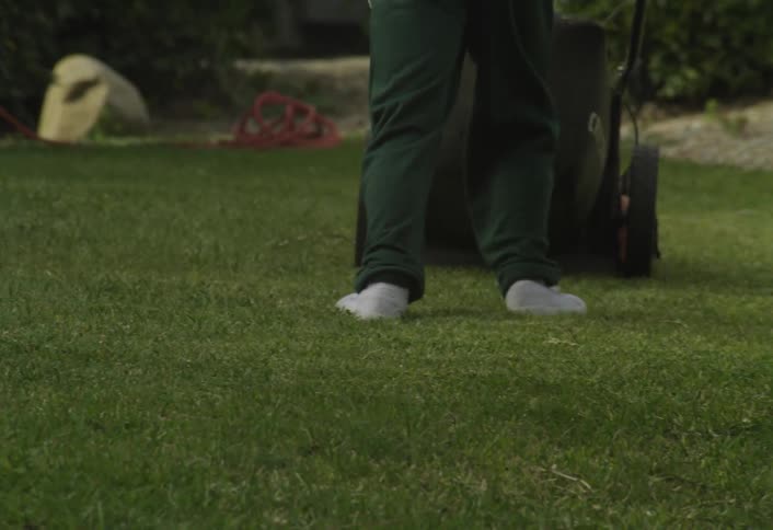 A child wearing a hat pushes a lawn mower across the backyard lawn at midday excitedly