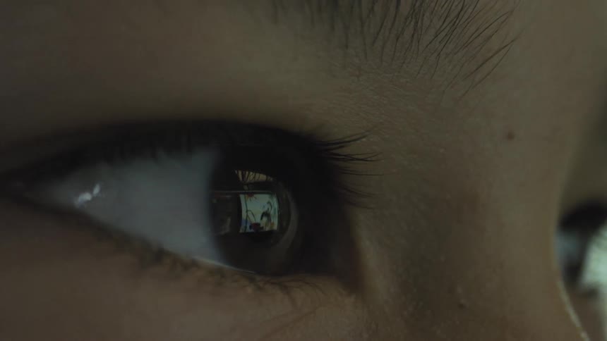 An extreme closeup of a persons eye in dim light shows the bright colorful reflection of a digital screen clearly visible in the pupil indicating the person is focused on watching media