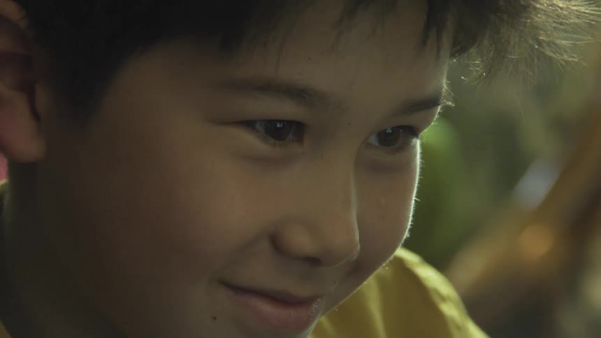 A closeup captures the gentle expression of a young boy in a yellow shirt as he gazes downward and breaks into a warm subtle smile under soft indoor lighting