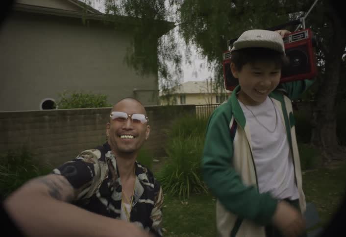 A man and a boy in a suburban yard both smiling at the camera and having fun the man gestures while the boy wearing a cap holds a red boombox on his shoulder
