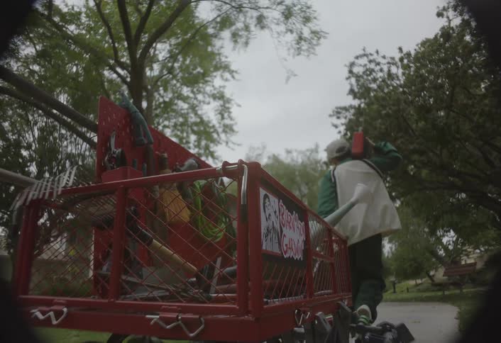 A low angle of a young boy wearing a coat and a baseball cap while holding a boombox with one hand pushing a red utility cart loaded with gardening tools down a path in a suburban setting