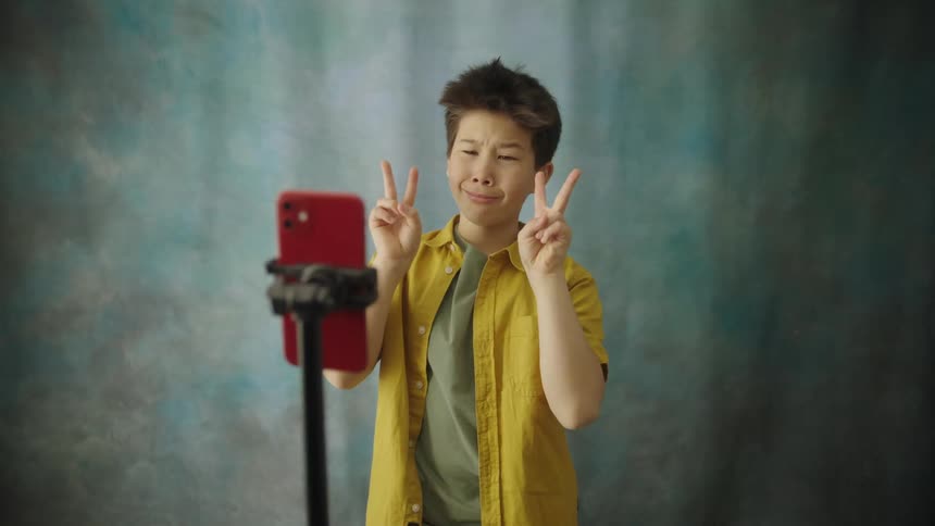 A young boy in a yellow shirt stands in front of a textured backdrop actively recording a video of himself using a red smartphone on a stand he poses makes silly faces and flashes a peace sign suggesting he is creating engaging content for social med