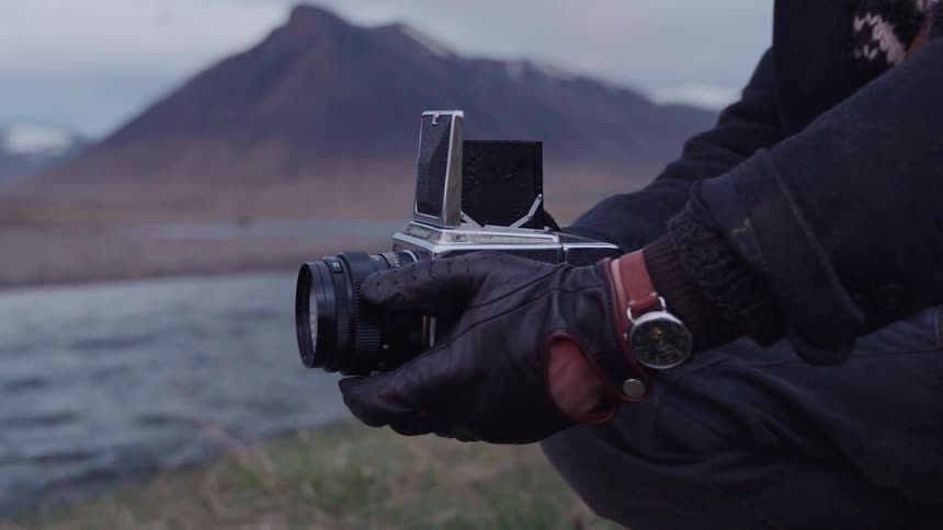 Clad in leather gloves a photographer holds a vintage mediumformat camera steady against a flowing stream and a vast dark mountain range in the distance