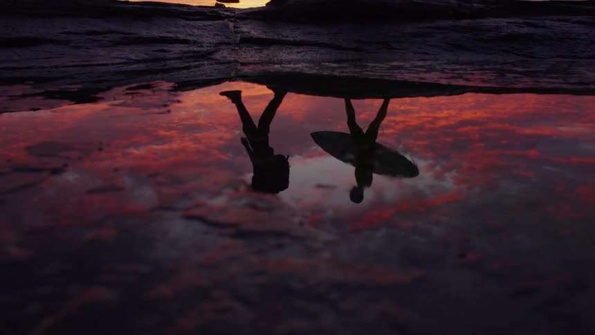 Reflection of two people walking with a surfboard in a tide pool at sunset