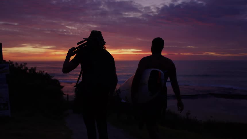 Silhouetted against a dramatic purple sunset a photographer and a surfer walk sidebyside along a coastal path carrying their gear as the ocean waves crash in the distance