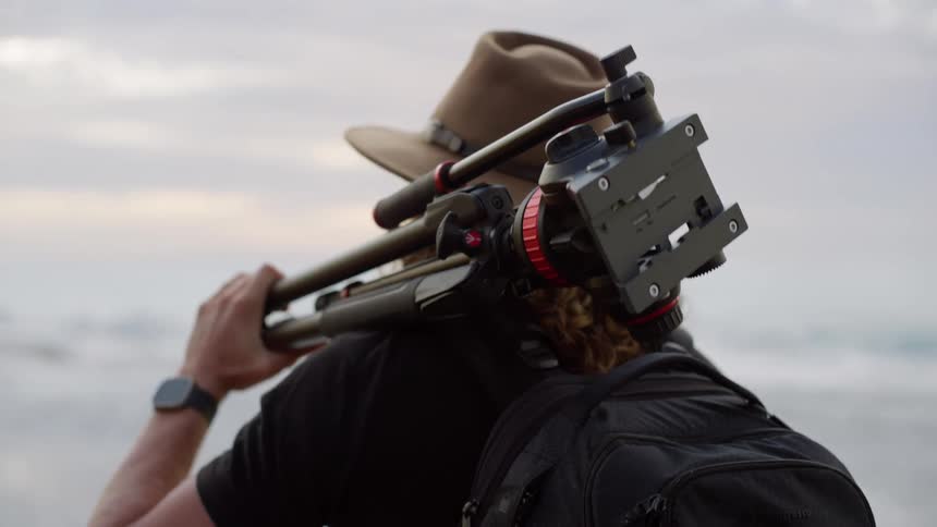 A man with curly hair and a hat carries a tripod over his shoulder looking forward on a beach