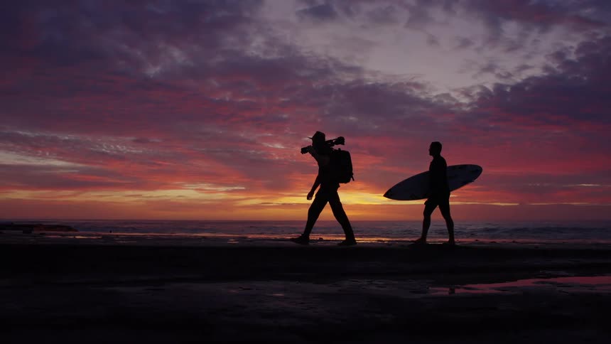 A surfer and a man with a camera walk along the beach as the sun is below the horizon