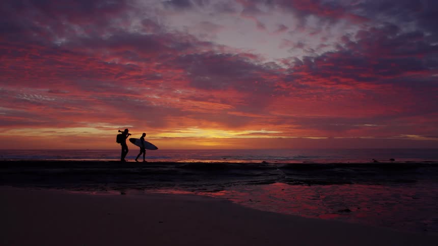 Two men walk on the beach as one man carries his surfboard and the other carries camera equipment under a colorful sunset sky