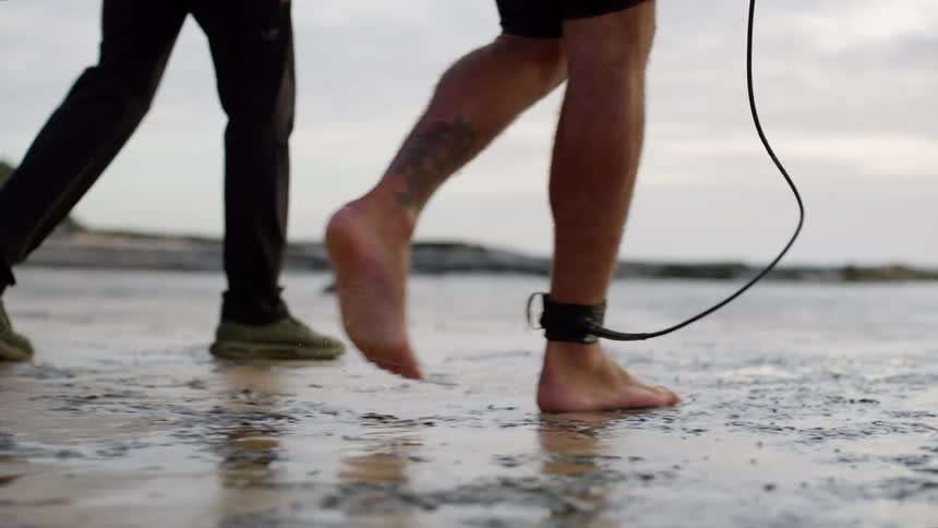 Two men walk on a wet beach one barefoot with an ankle surf leash heading toward the water