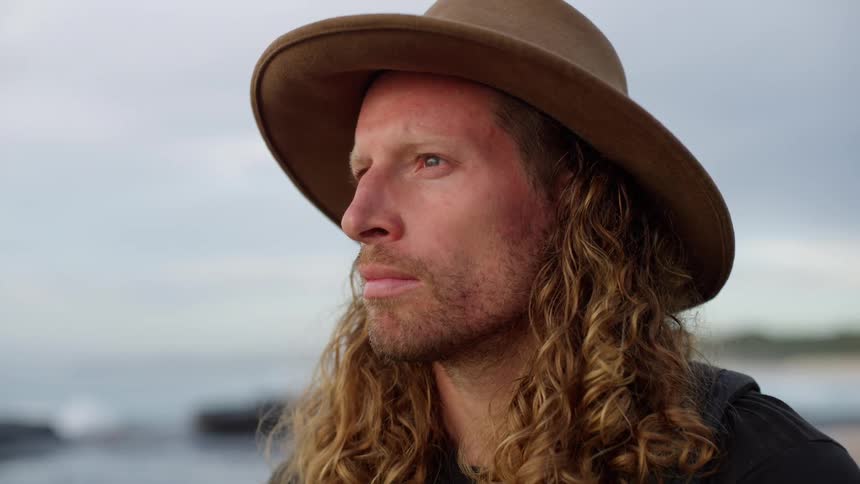 A closeup side profile focuses on a man wearing a widebrimmed hat showcasing his long curly hair and beard he looks offcamera with a contemplative expression against a soft overcast backdrop of a rocky shoreline