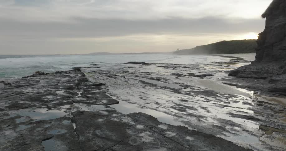 This is a wide static shot capturing a rugged cloudy coastlinethe foreground is dominated by dark flat and wet rock formations leading towards the crashing waves of the ocean with a steep rocky cliff visible on the right