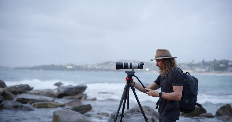 A photographer wearing a hat and backpack stands beside his camera on a tripod along a rocky coastline