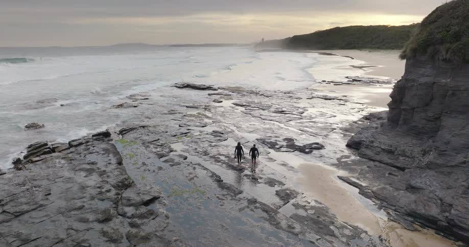 Two men walk across wet coastal rocks along a wide overcast shoreline at dusk