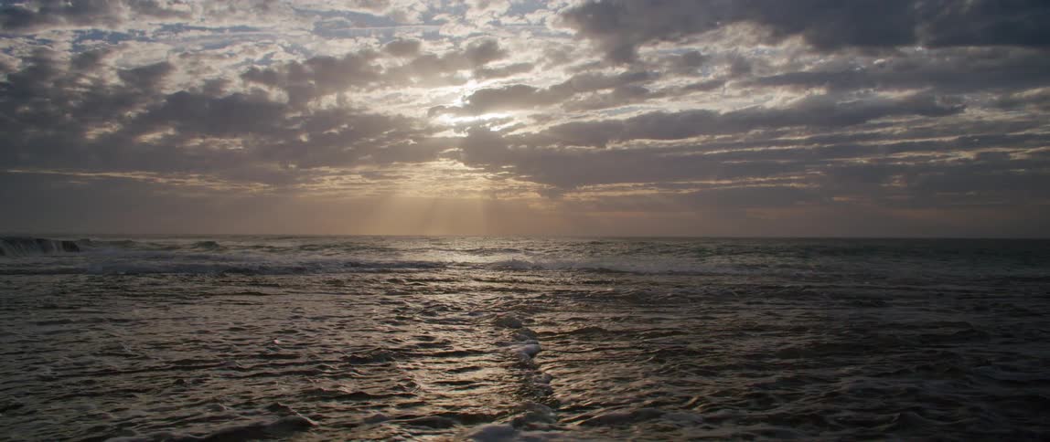 Waves roll in to shore and over the beach under a cloudy sky